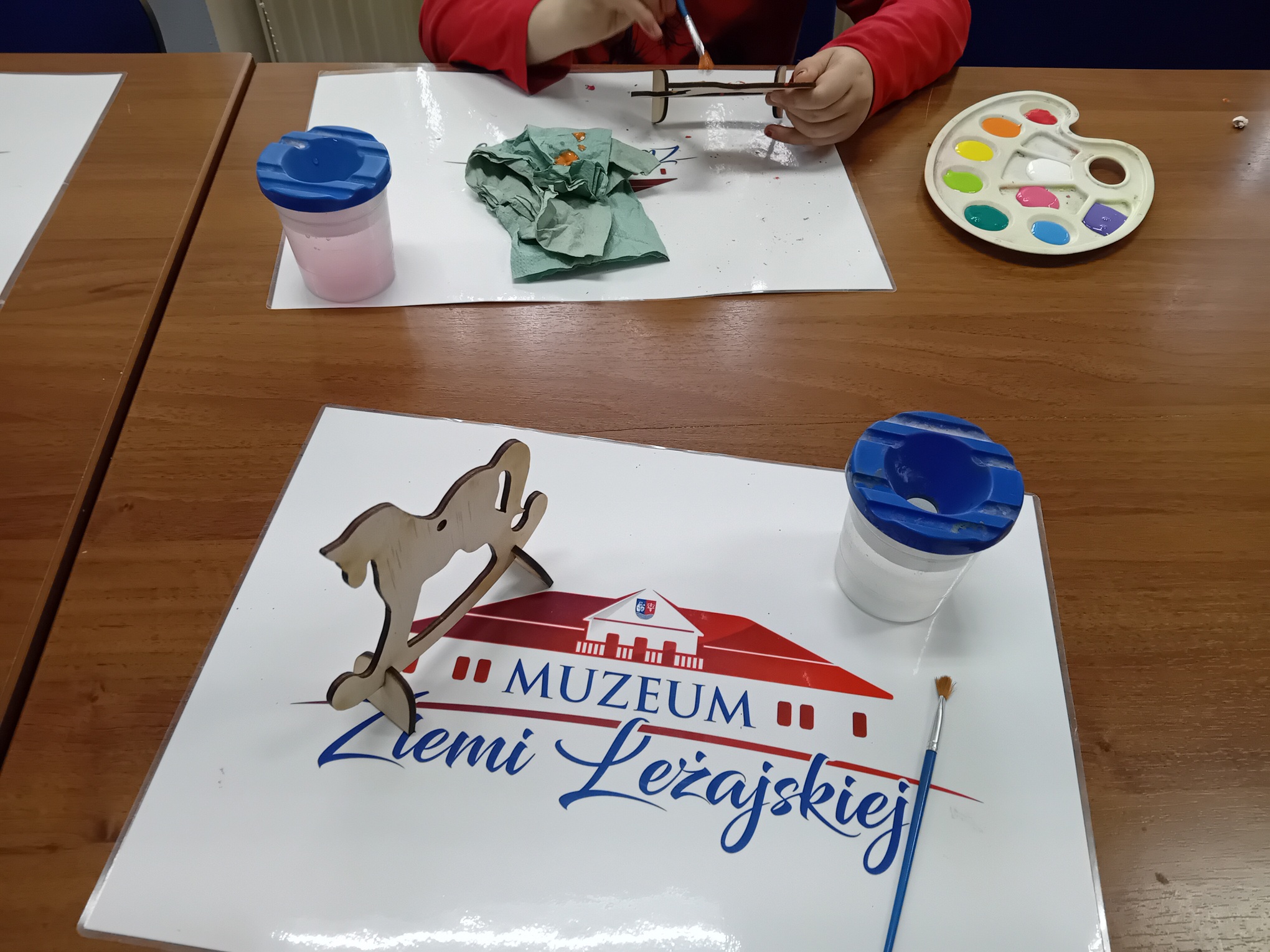 Dziecko malujące drewnianą figurkę konia na biegunach. Na stole leży paleta z&nbsp;farbami i&nbsp;podkładka z&nbsp;napisem Muzeum Ziemi Leżajskiej.
