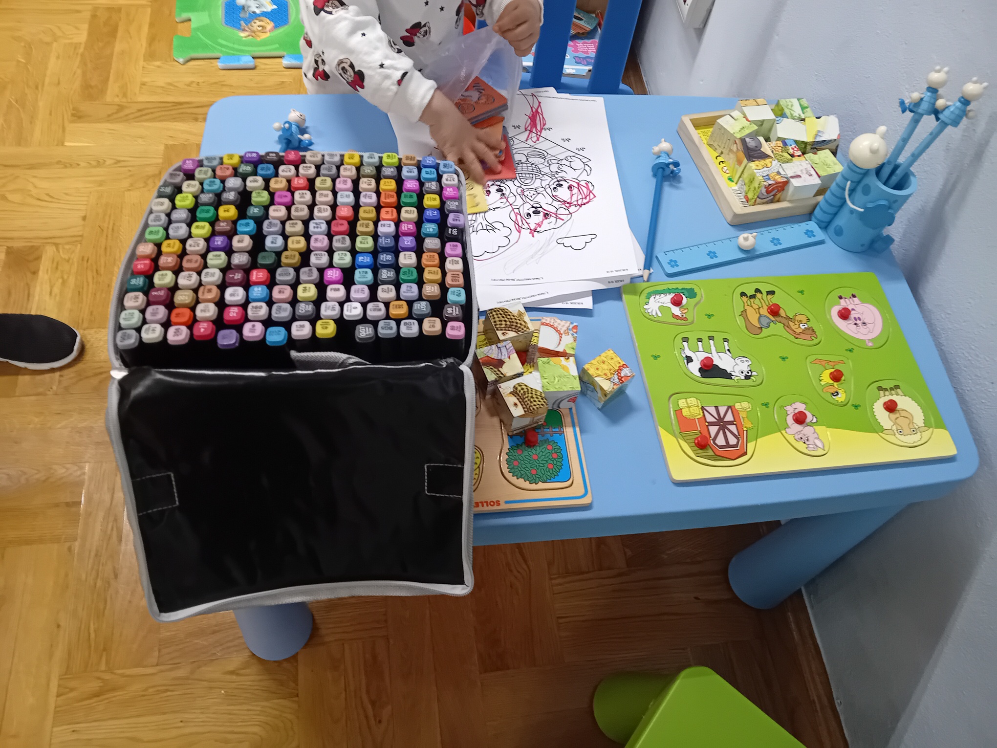 Niebieski stolik dziecięcy, na którym znajduje się duży zestaw kolorowych markerów, drewniane puzzle ze zwierzętami i&nbsp;kolorowanki.