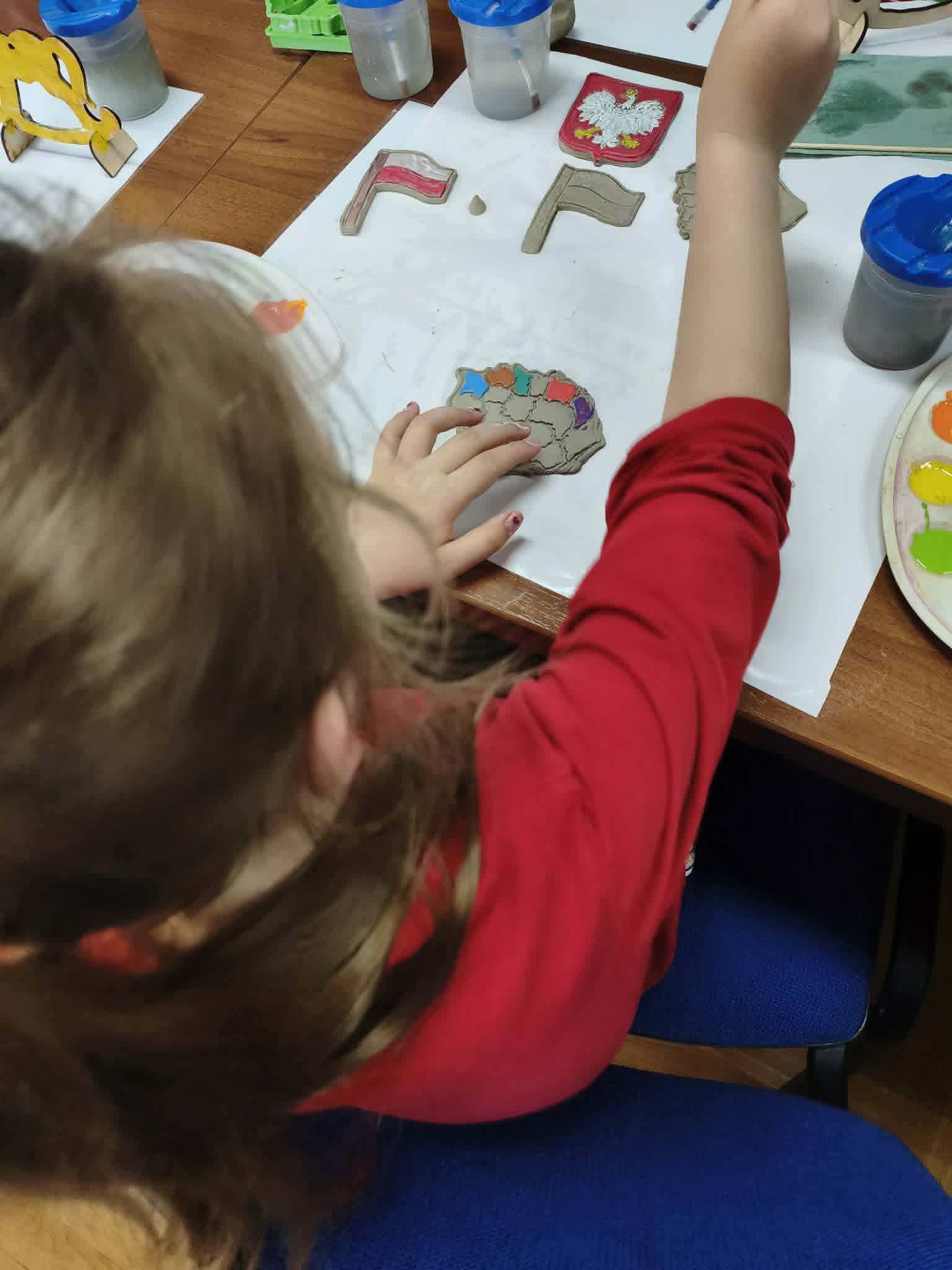Dziecko maluje farbami gliniany model mapy Polski. Na stole stoją kubki z&nbsp;wodą, paleta z&nbsp;farbami i&nbsp;inne patriotyczne figurki.